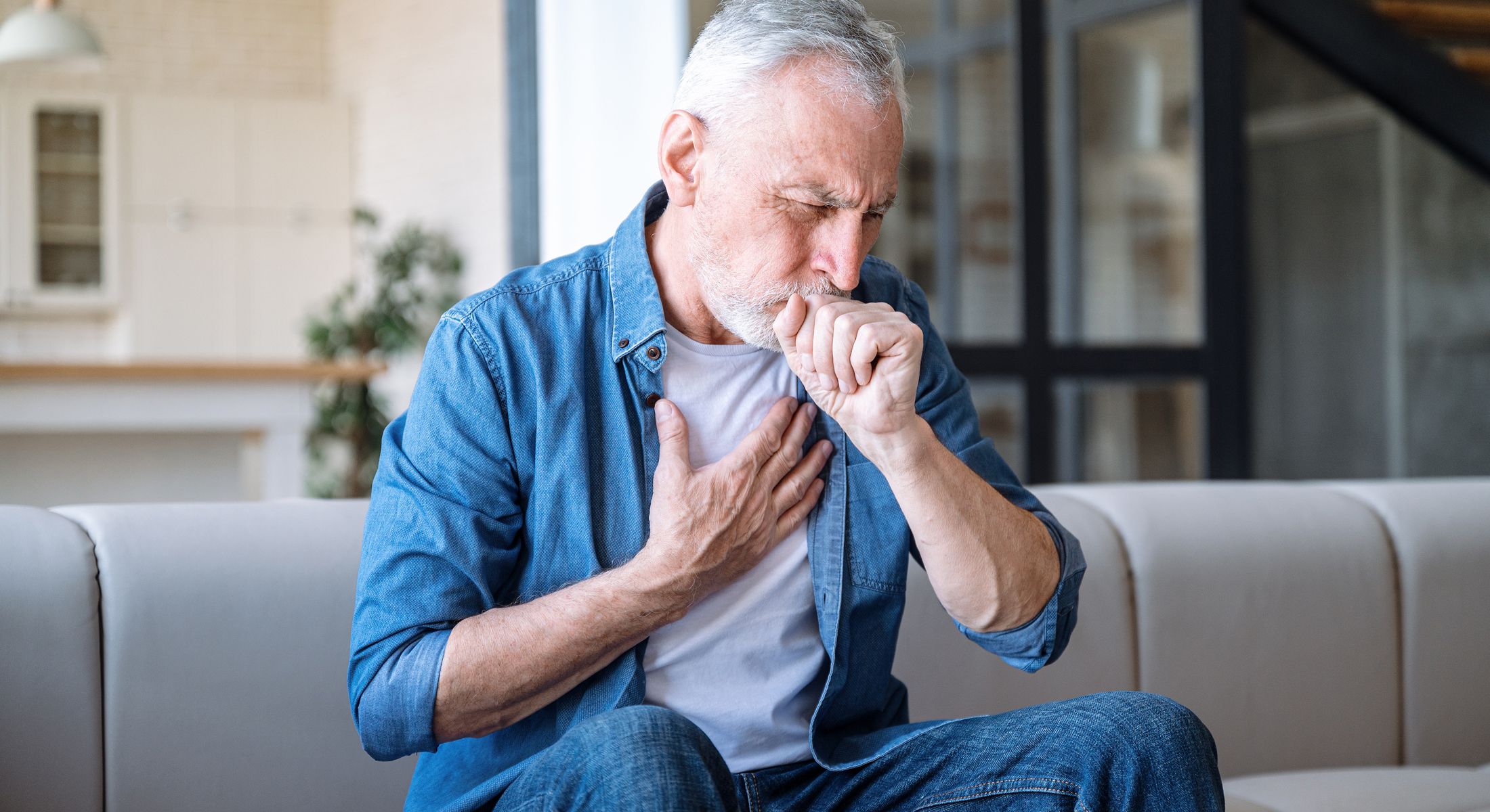 Elderly man coughing at home, looking concerned.